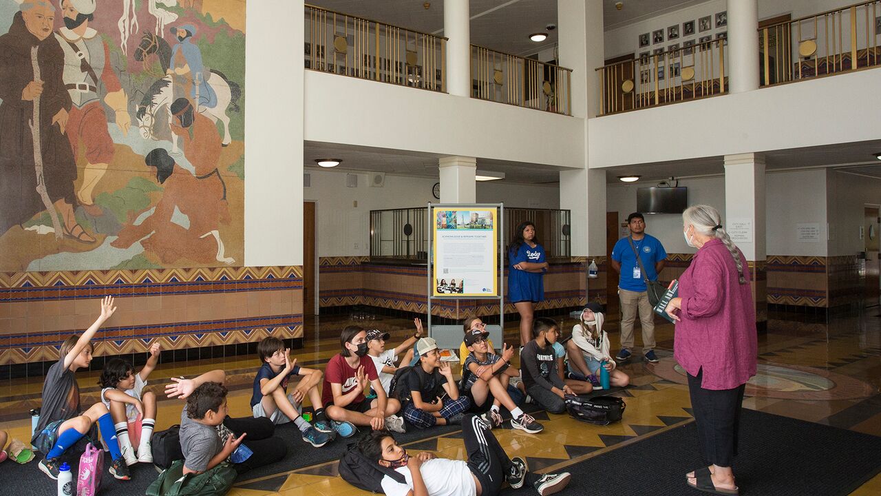 School kids sitting on the floor in historic city hall by mural listening to artist Glenna Avila