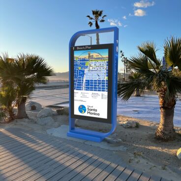 Digital Kiosk in a Blue Frame with an interactive map showing on the screen - the kiosk is located next to a beach walking path and is surrounded by palm trees