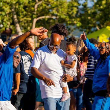 An adult holds a toddler and dance with a group at the Juneteenth Celebration at Virginia Avenue Park