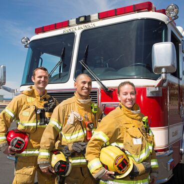 Three Santa Monica Fire Department Firefighters in yellow turnouts, holding their helmets standing in front of a fire engine at the beach