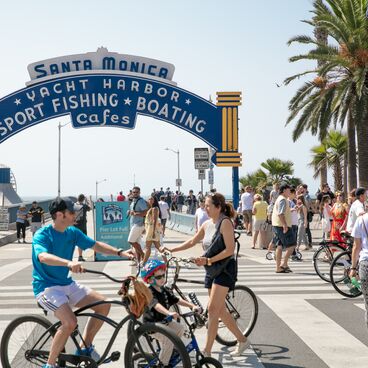 Group of People Walking and Riding Bikes in front of the Santa Monica Pier Sign
